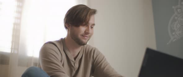 A close up of a young man smiling while sitting in bed and working on a computer