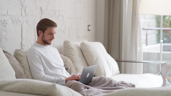 Cheerful Caucasian Man Sits at Home on the Couch and Works Using a Laptop Financial Manager on a