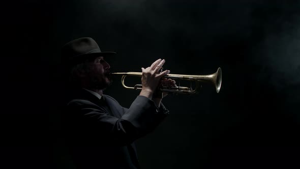 Jazzman in Jacket and Hat Plays Trumpet on Dark Background in the Fog Side View