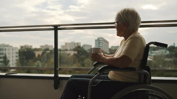 Old Pensive Disabled Woman in Wheelchair on the Terrace Drinking a Cup of Tea or Coffee. 