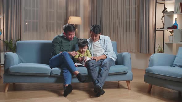 Modern Married Gay Couple Spending Time With Their Son Sitting On Sofa In Living Room Reading Book