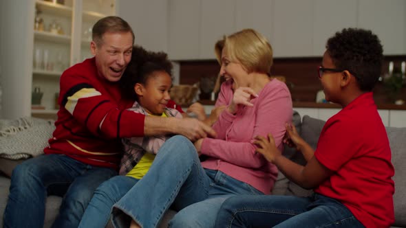 Excited School Age Multiracial Kids and Grandparents Having Fun Tickling on Sofa