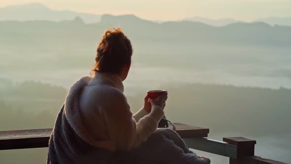 Young Woman is Sitting with a Mug of Coffee on a Cliff Overlooking the Mountains with Fog