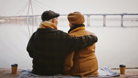 Aged Couple are Hugging Sitting on Bench Carved From Tree Trunk on Bank of a River and Looking at