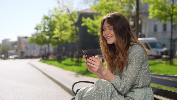 Portrait of Curly Young Woman 20s Wearing Dress Smiling and Holding Smartphone While Sitting on