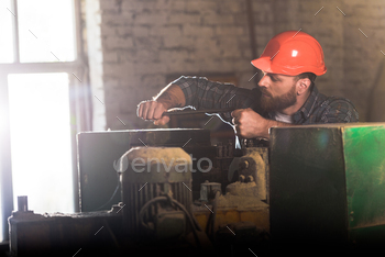 bearded worker in protective helmet repairing machine tool at sawmill