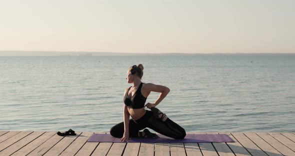 Woman Is Practicing Yoga and Doing Pigeon Pose on Mat on River Pier, Side View.