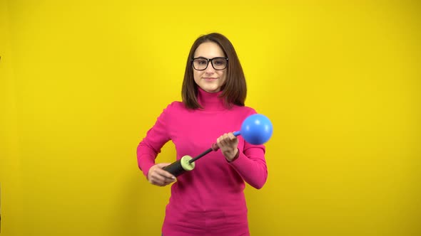 A Young Woman Inflates a Blue Balloon with a Pump on a Yellow Background. Girl in a Pink Turtleneck