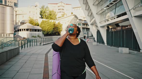 Tired After Sport Training Overweight African American Woman Going Home with Fitness Mat Drinking