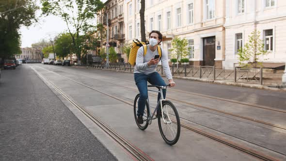 Courier with Yellow Backpack and Protective Mask Rides a Bicycle on the Street with Food Delivery