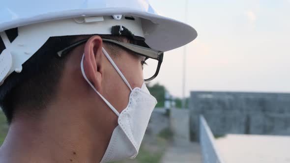 Young Asian engineer wearing a helmet and mask looks and smiles at the camera on the dam background.