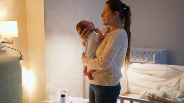 Happy Smiling Young Mother Cuddling and Holding Her Newborn Baby in Bedroom at Night