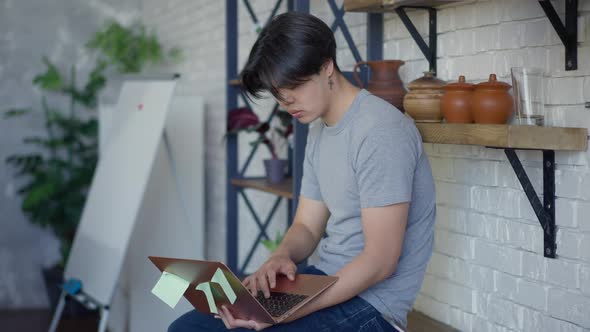 Side View of Asian Young Man Messaging Online Typing on Laptop Keyboard Sitting on Kitchen