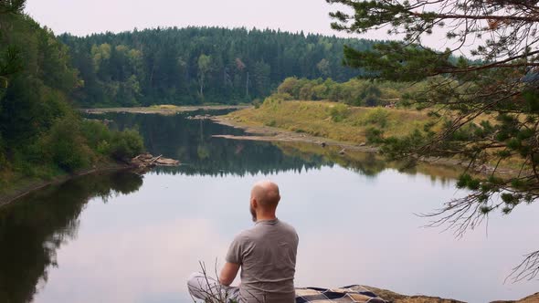 A Bald Man with a Beard Sits on the Edge of a River Bank Cliff and Meditates in a Calm Environment