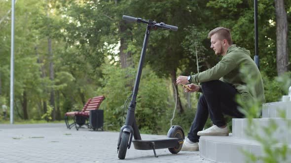 Man is Sitting on Stairs in Park and Typing Something on Phone Next to Him is an Electric Scooter