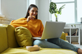 Attractive young woman using laptop and enjoying cookies while sitting on the couch at home - PhotoDune Item for Sale