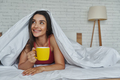 Dreamful woman looking out of a blanket and holding coffee cup while lying in bed at home - PhotoDune Item for Sale
