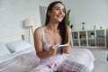 Beautiful young woman holding pregnancy test and smiling while sitting on bed at home - PhotoDune Item for Sale