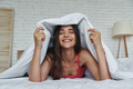 Beautiful young woman covering with blanket and smiling while lying in bed at home - PhotoDune Item for Sale