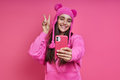 Beautiful young woman in funky hat making selfie and gesturing against pink background - PhotoDune Item for Sale