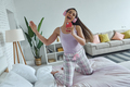 Young woman using smart phone like microphone while dancing on bed at home - PhotoDune Item for Sale