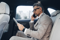 Confident mature businessman using digital tablet and talking on phone while sitting in the car - PhotoDune Item for Sale