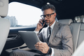 Confident businessman using digital tablet and talking on phone while sitting in the car - PhotoDune Item for Sale