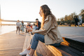 Young casual woman in coat sitting on bench on sea embankment using smartphone - PhotoDune Item for Sale