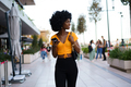Smiling afro-american woman holding mobile phone while walking in the street - PhotoDune Item for Sale