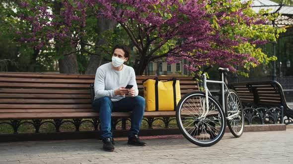 Caucasian Delivery Man Sitting on Bench in Central Park and Using Smartphone with Yellow Backpack