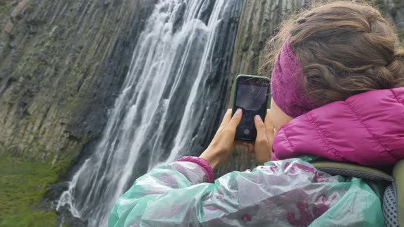 Lady Holds Phone and Takes Photo Standing Near Waterfall