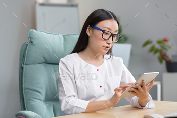 l Asian woman doctor records a patient’s medical history online in a tablet. Typing in a tablet. Sitting at the table in the winding office.