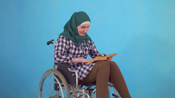 Young Muslim Woman Disabled in Wheelchair with Book in Hand on Blue Background