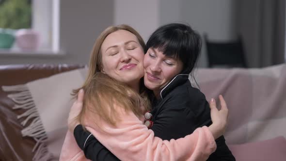 Two Happy Women Hugging Smiling Looking at Camera Sitting in Comfortable Living Room on Cozy Couch