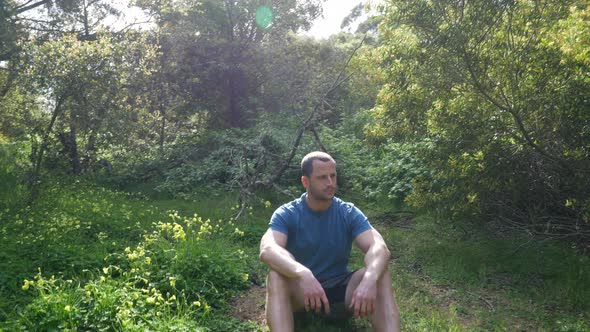 A handsome man sitting in a green grassy forest meadow on a bright sunny day while thinking and pond