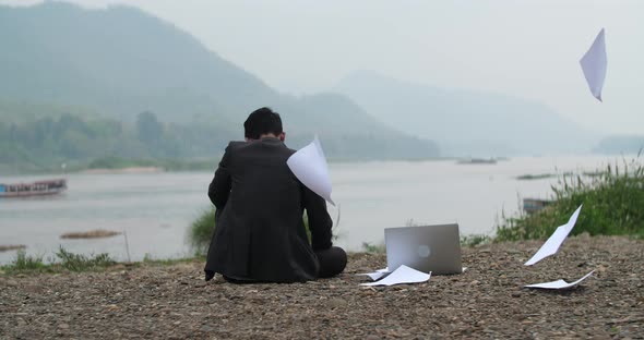 Businessman Sitting Near River With Computer And Throwing Papers