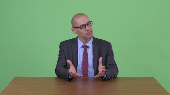 Happy Bald Multi Ethnic Businessman Presenting While Holding Clipboard Behind Desk