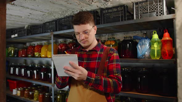 Satisfied Happy Young Farmer in Glasses Holding Notebook and Counting Profits in Basement