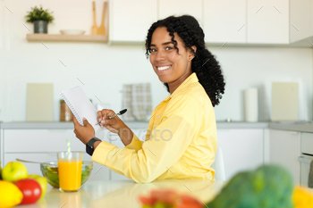 ration diet or menu in notebook, sitting near fresh salad and juice at kitchen table, looking and smiling at camera