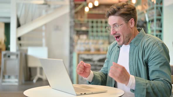 Excited Middle Aged Man Celebrating Success on Laptop in Cafe