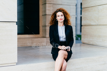 Attractive young woman wearing black formal suit, sitting crossed legs with electronic device