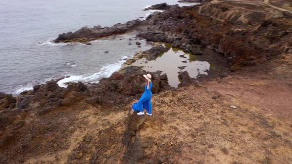 Aerial View of Woman in a Beautiful Blue Dress and Hat Stands on Top of a Mountain in a Conservation