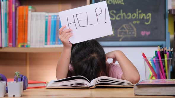 Asian little girl feeling sad and tired sitting at the table with many books and holding a paper.