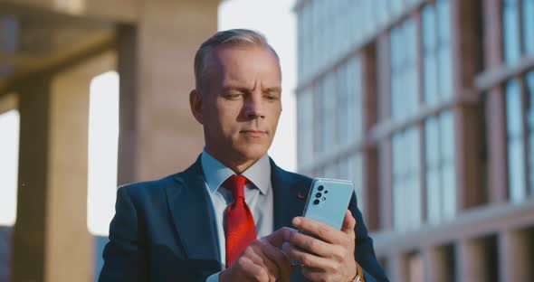 Serious Businessman Looking at Cellphone Typing Message Standing Outdoors Downtown