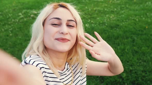 Picture of a Young Blond Cheerful Girl Standing in the Park and Taking a Selfie By Waving Medium