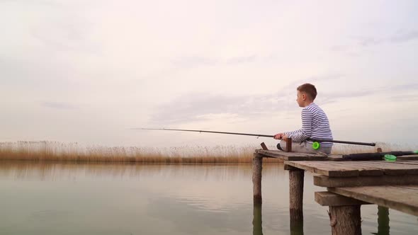 A Boy Sits on a Wooden Bridge and Catches a Fish on Fishing Rod