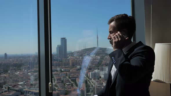 Young Businessman in Office Suit Is Talking Phone Near the Window with Panoramic City View.