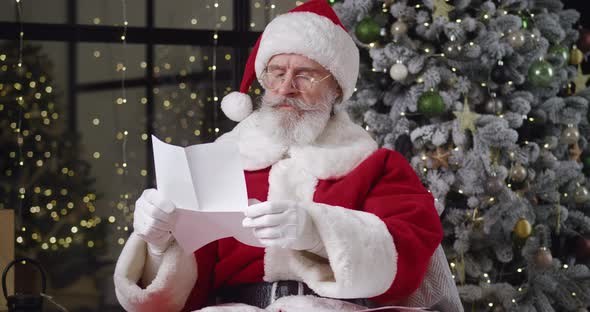 Front View of Cheerful Santa Claus Wearing Christmas Costume with Red Hat Sitting in Armchair