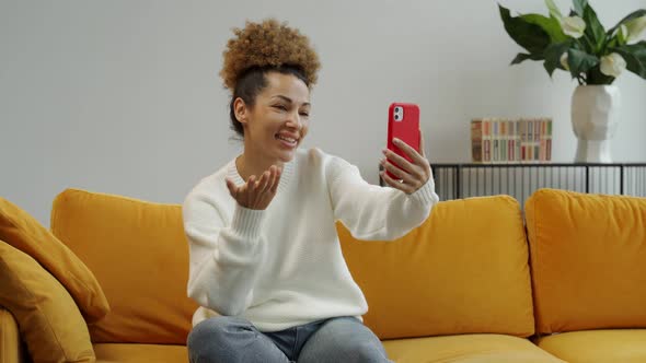 A Happy AfricanAmerican Woman with a Smartphone Makes a Video Call at Home Sitting on a Yellow Sofa