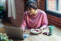 Beautiful young woman working with laptop while eating yogurt with fruits bowl at home. - PhotoDune Item for Sale
